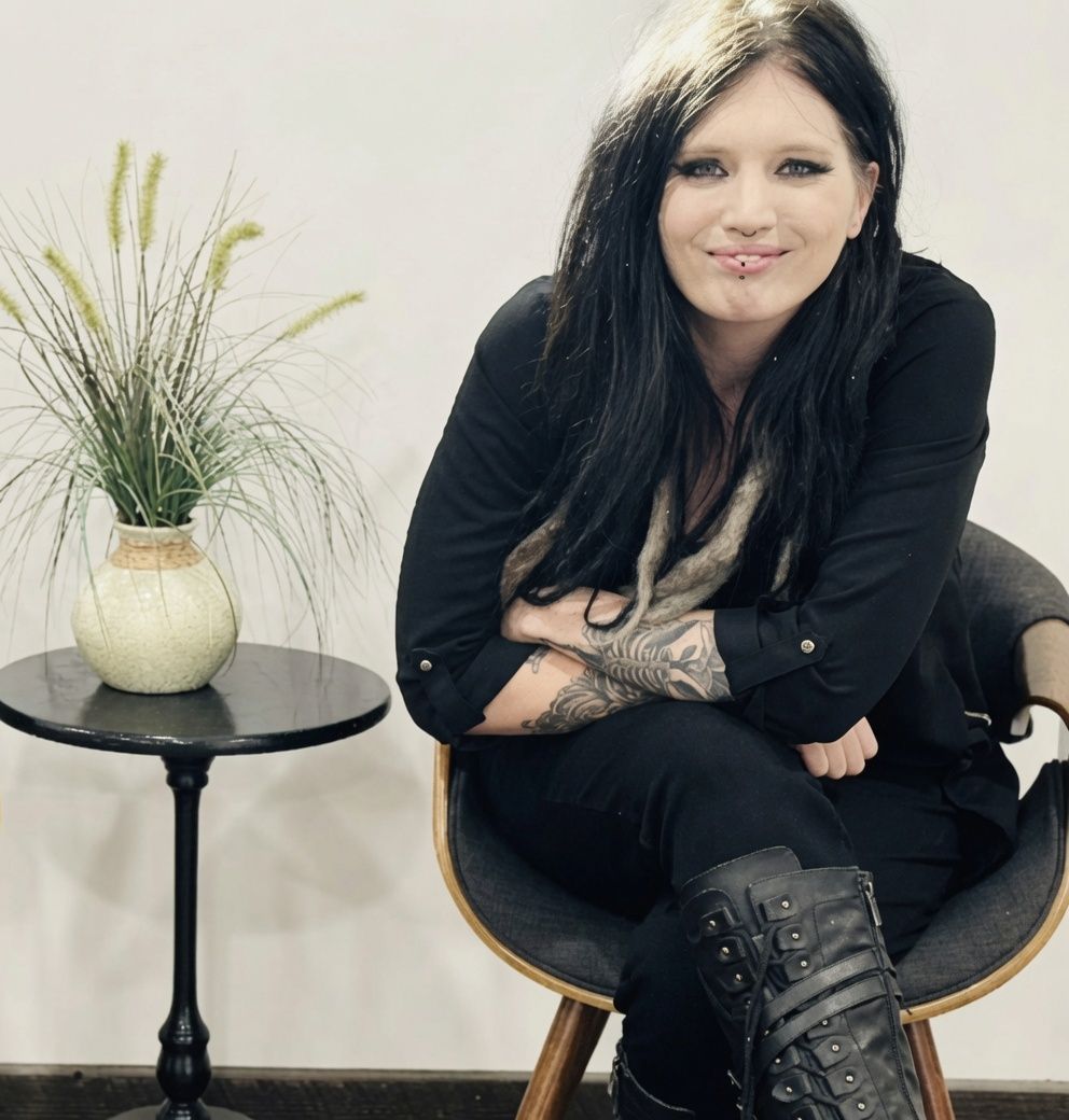 woman sitting in chair with black hair and all black clothes, next to a table with a plant