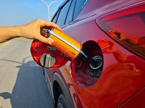 Hand pouring liquid from orange container into a red car's gas tank. Outdoors, sunny.