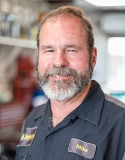 Man with a graying beard in a mechanic's uniform smiling, with a
