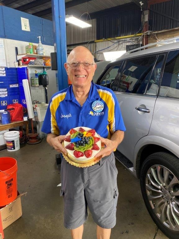 Man holding cake with fruit in a car repair shop; smiling near a silver SUV.
