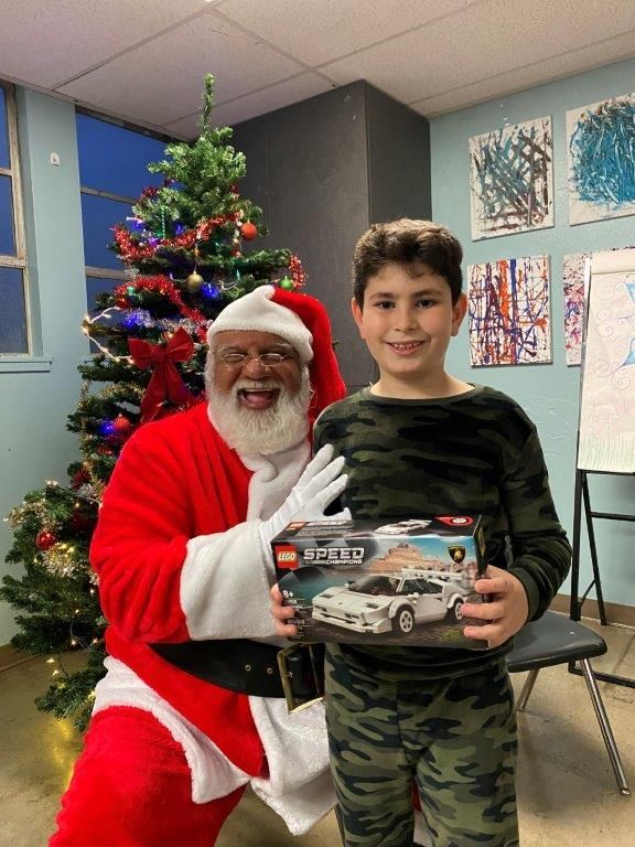 Santa Claus and a boy smiling together, holding a LEGO car set, next to a Christmas tree.
