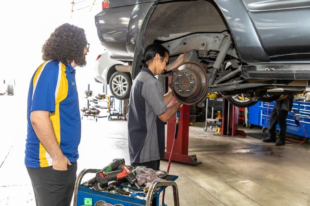 Mechanic working on a car, another watching. Workshop setting. Tools, lift, and other vehicles present.