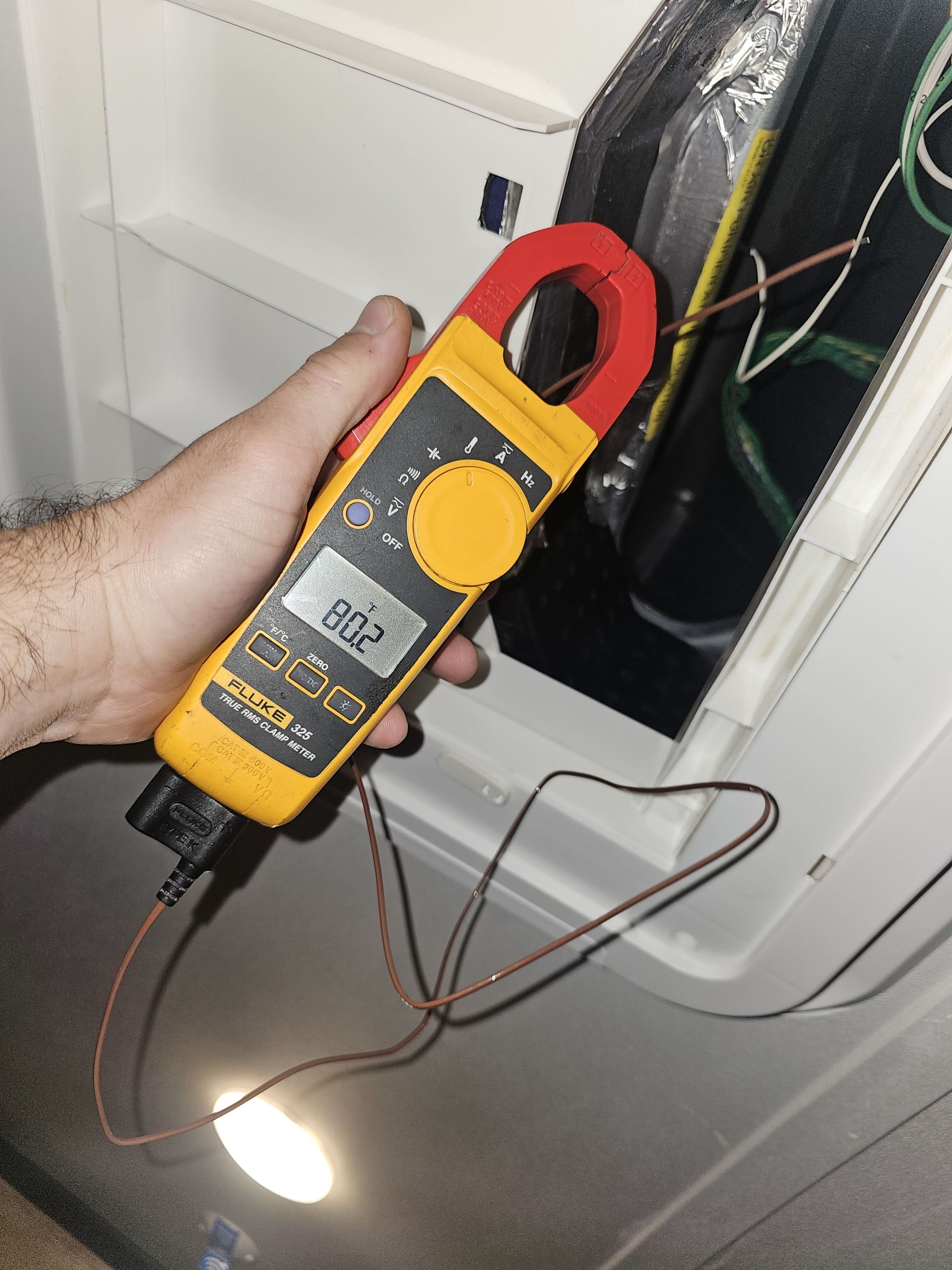 Hand holding a yellow clamp meter near an open electrical panel with exposed wiring and a lit flashlight below