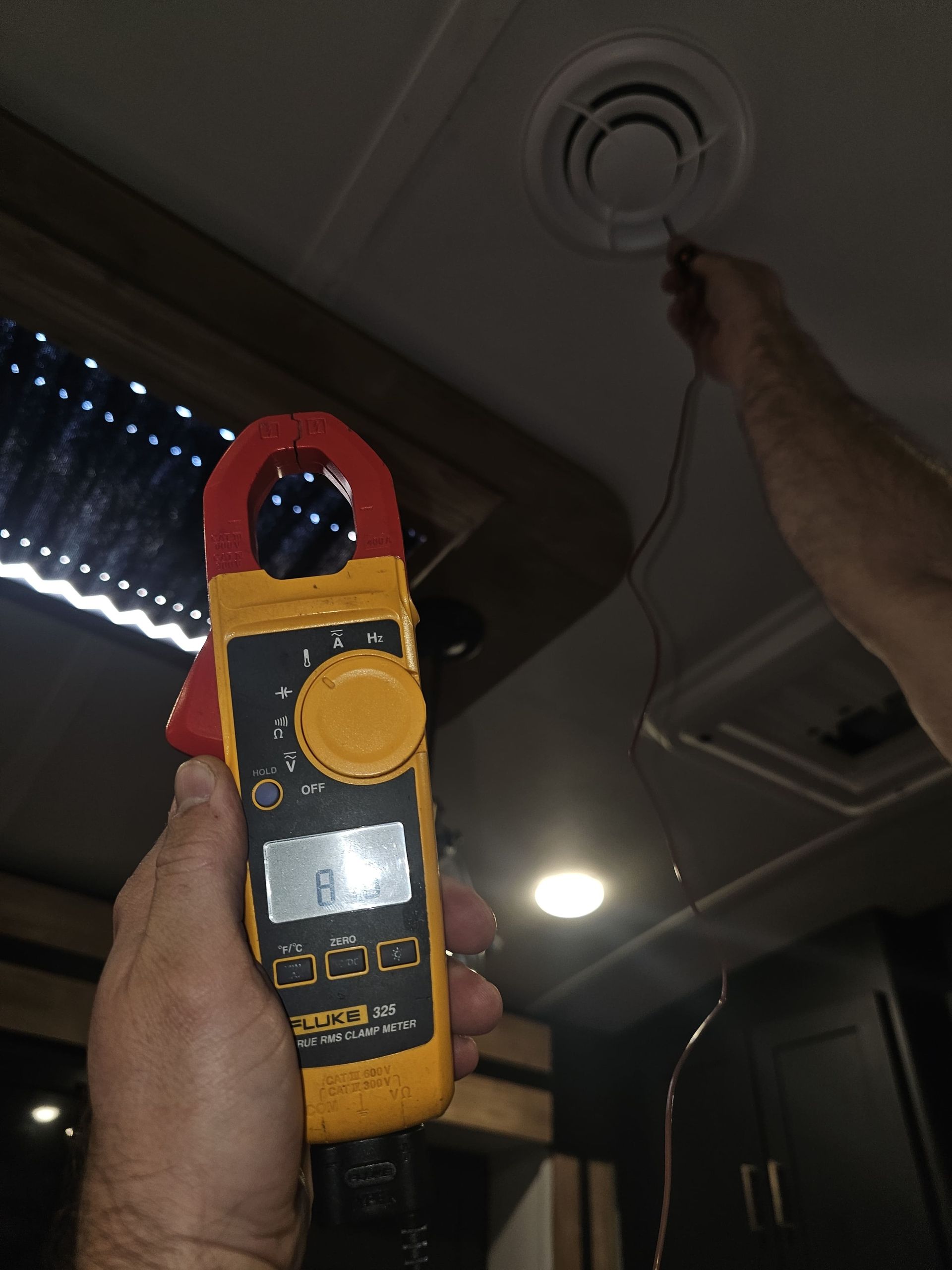 Person holding a yellow-and-red electric meter toward a ceiling smoke detector in a dim room