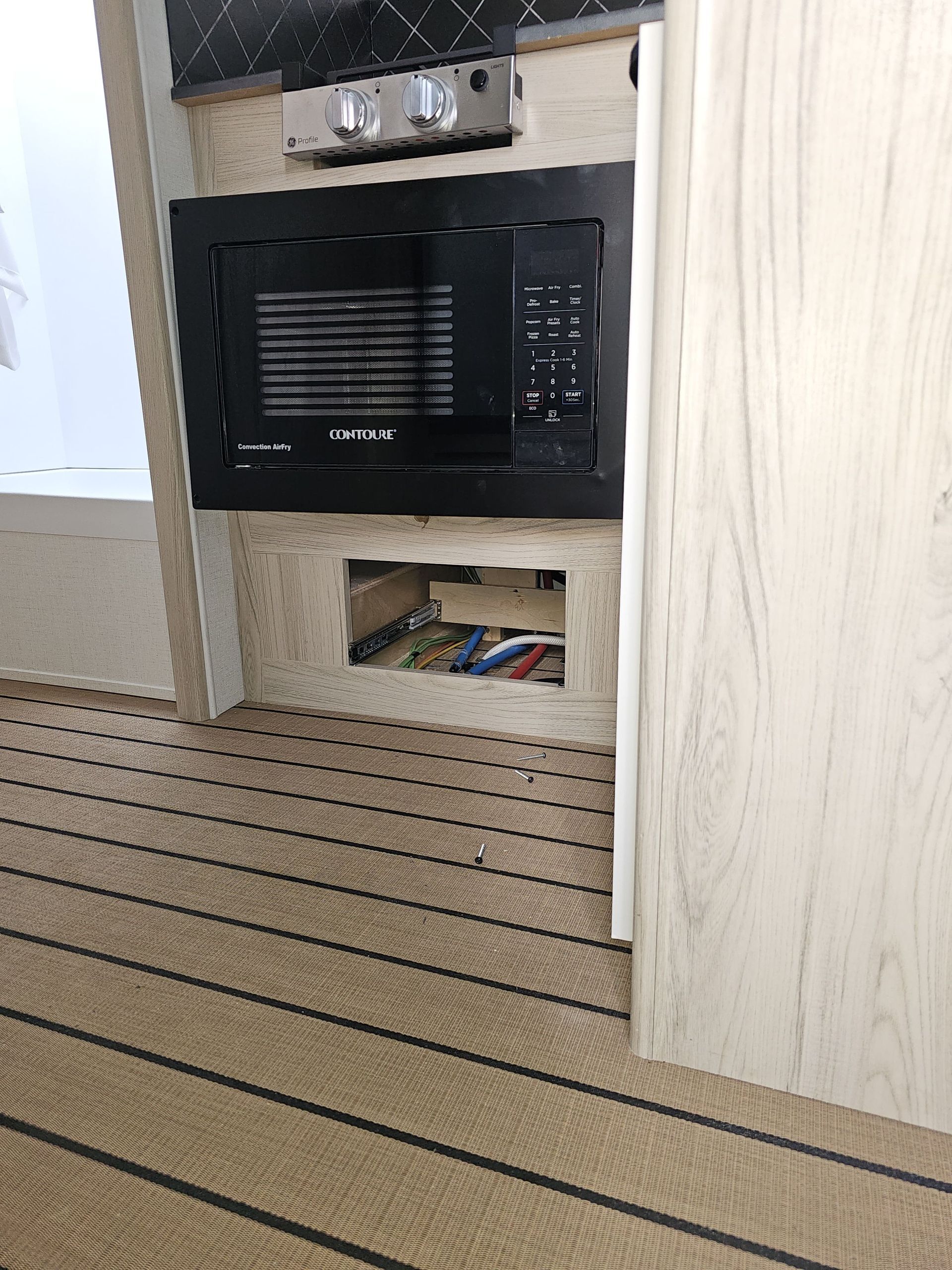 Built-in black microwave in a beige cabin wall with a small storage opening below, beside a striped carpet.