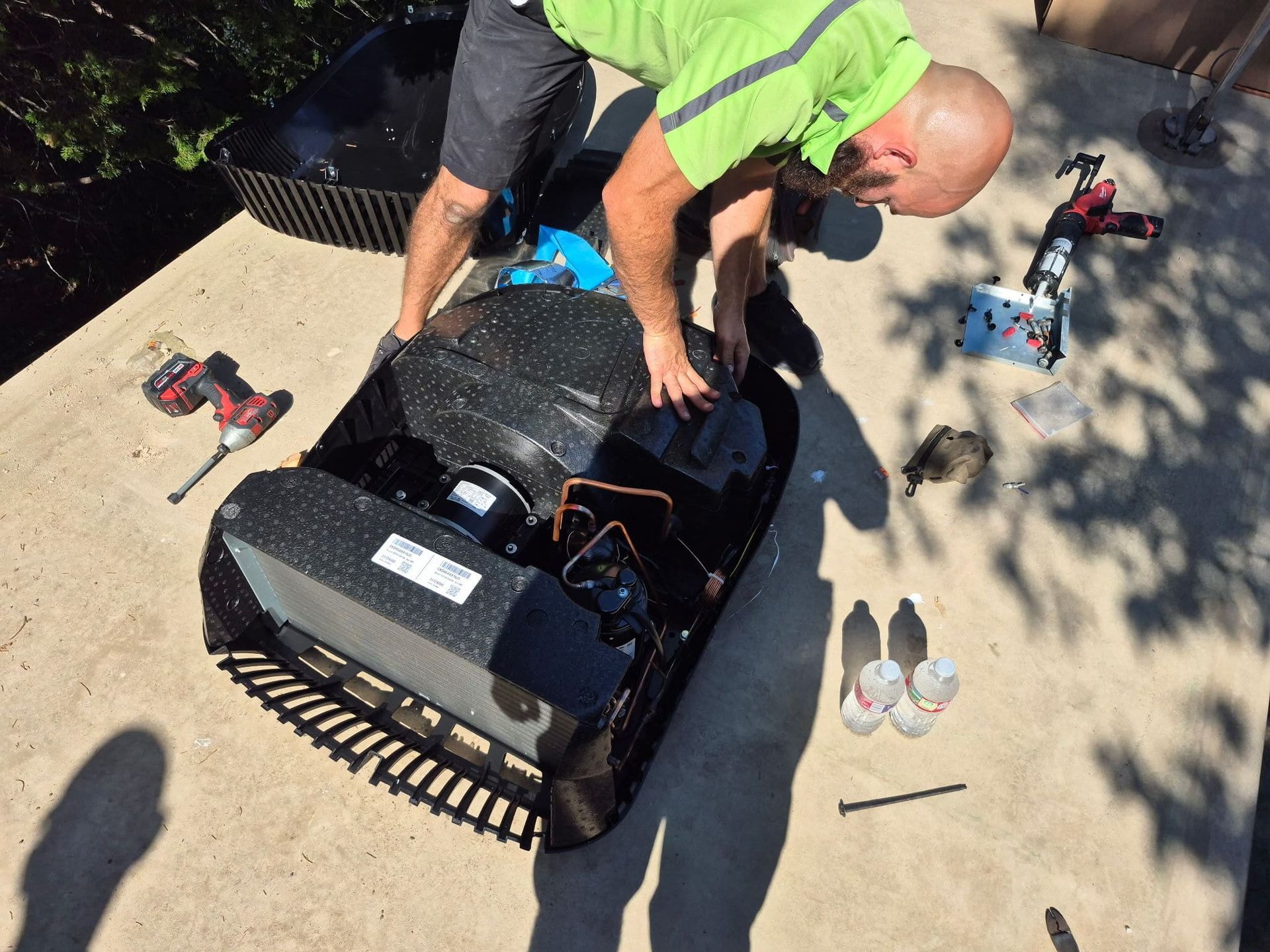 Worker kneels over a small tracked robot on pavement, with tools and parts scattered nearby.