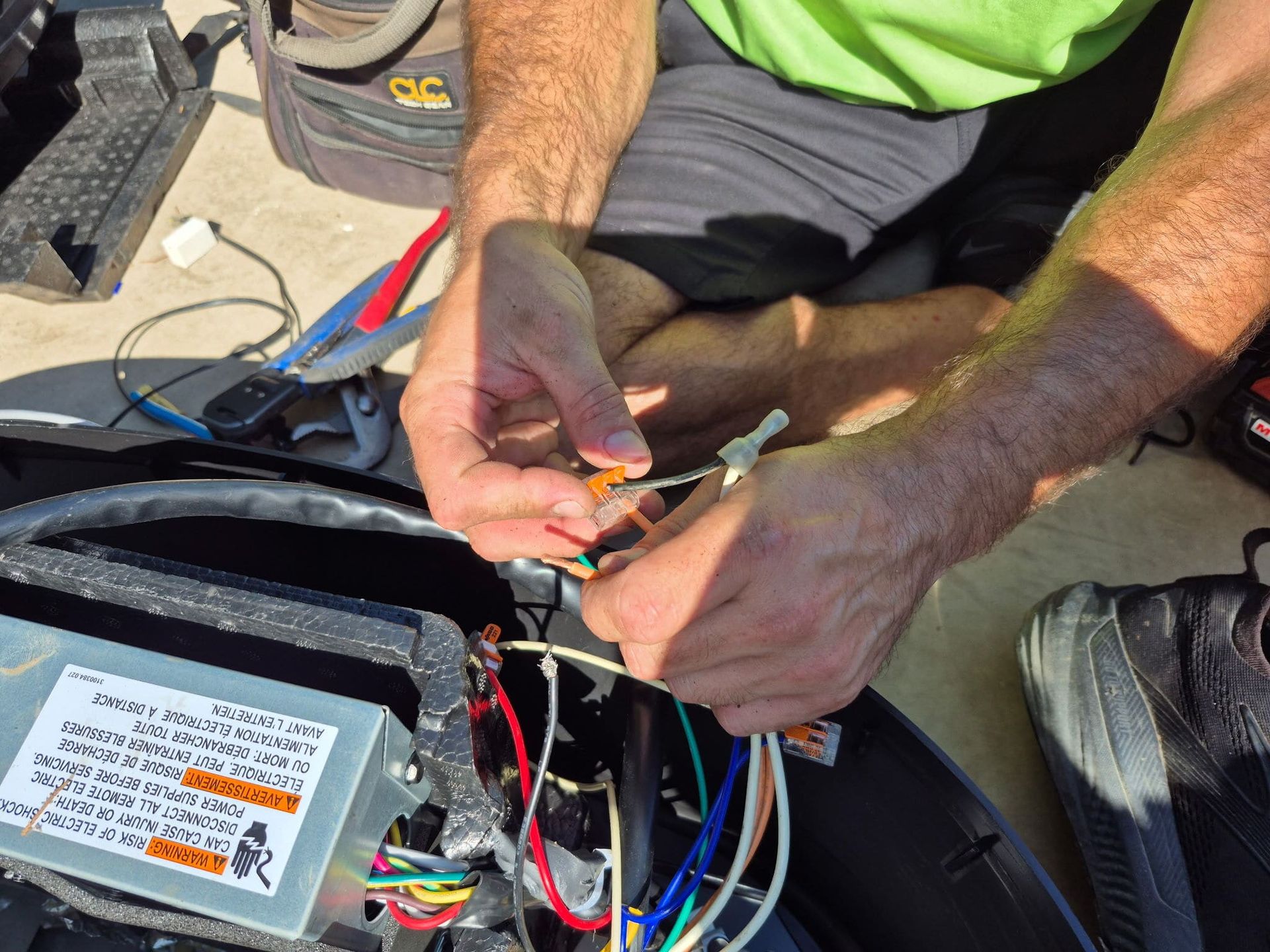 Person wiring cables inside an open device on the ground, with tools nearby and a label on the unit
