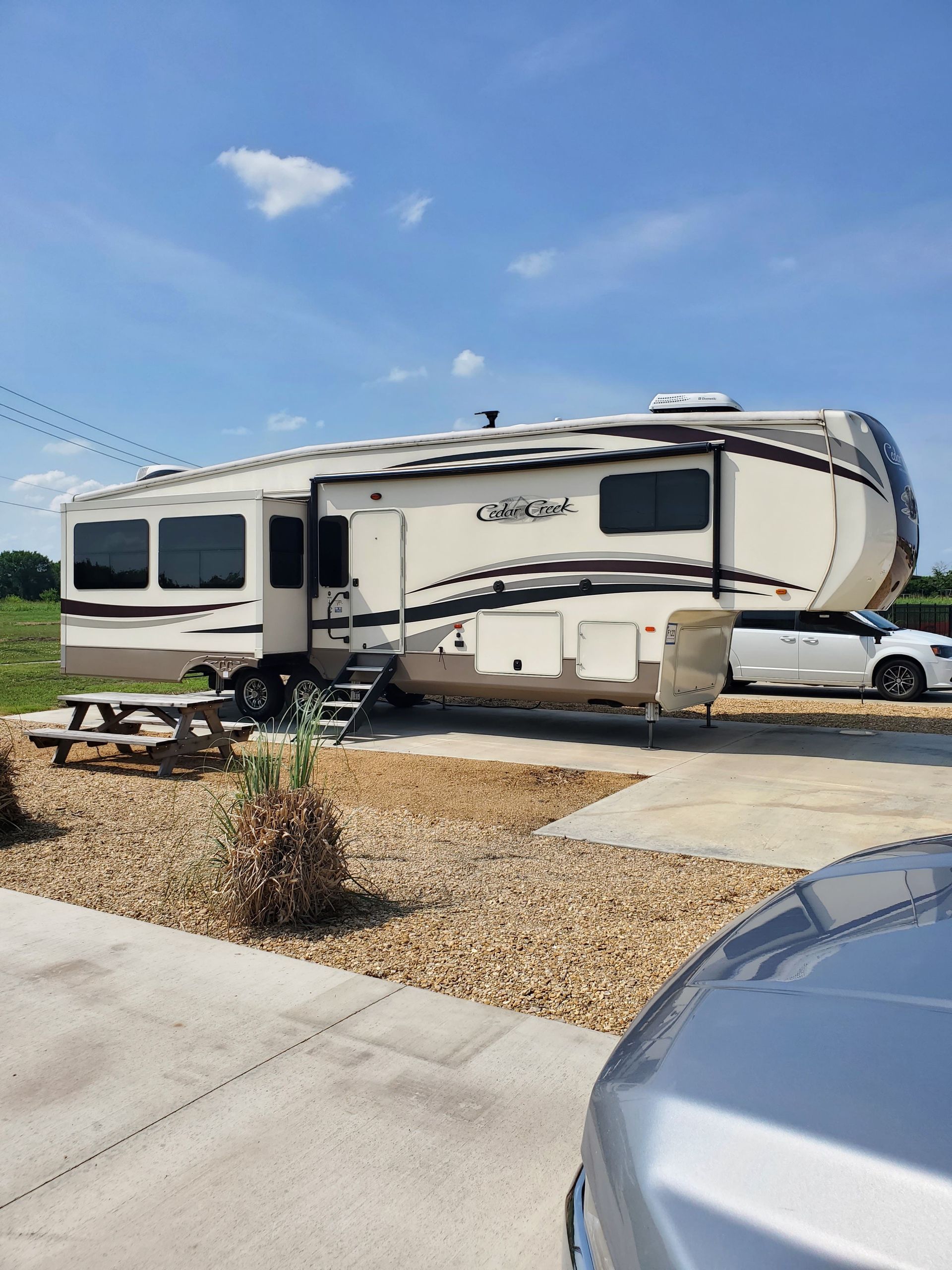 Large beige RV parked in a residential driveway under a blue sky, with a car beside it.