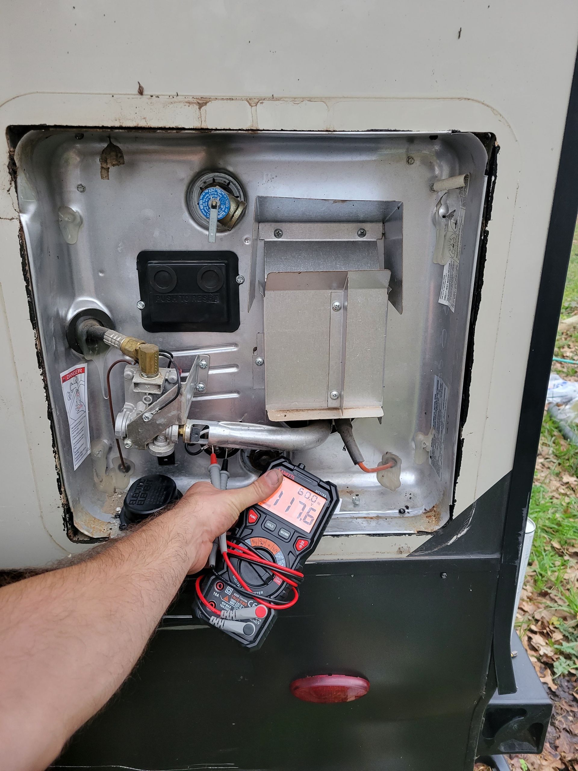 Hand holding a multimeter near an open utility meter box with exposed wiring and metal components.