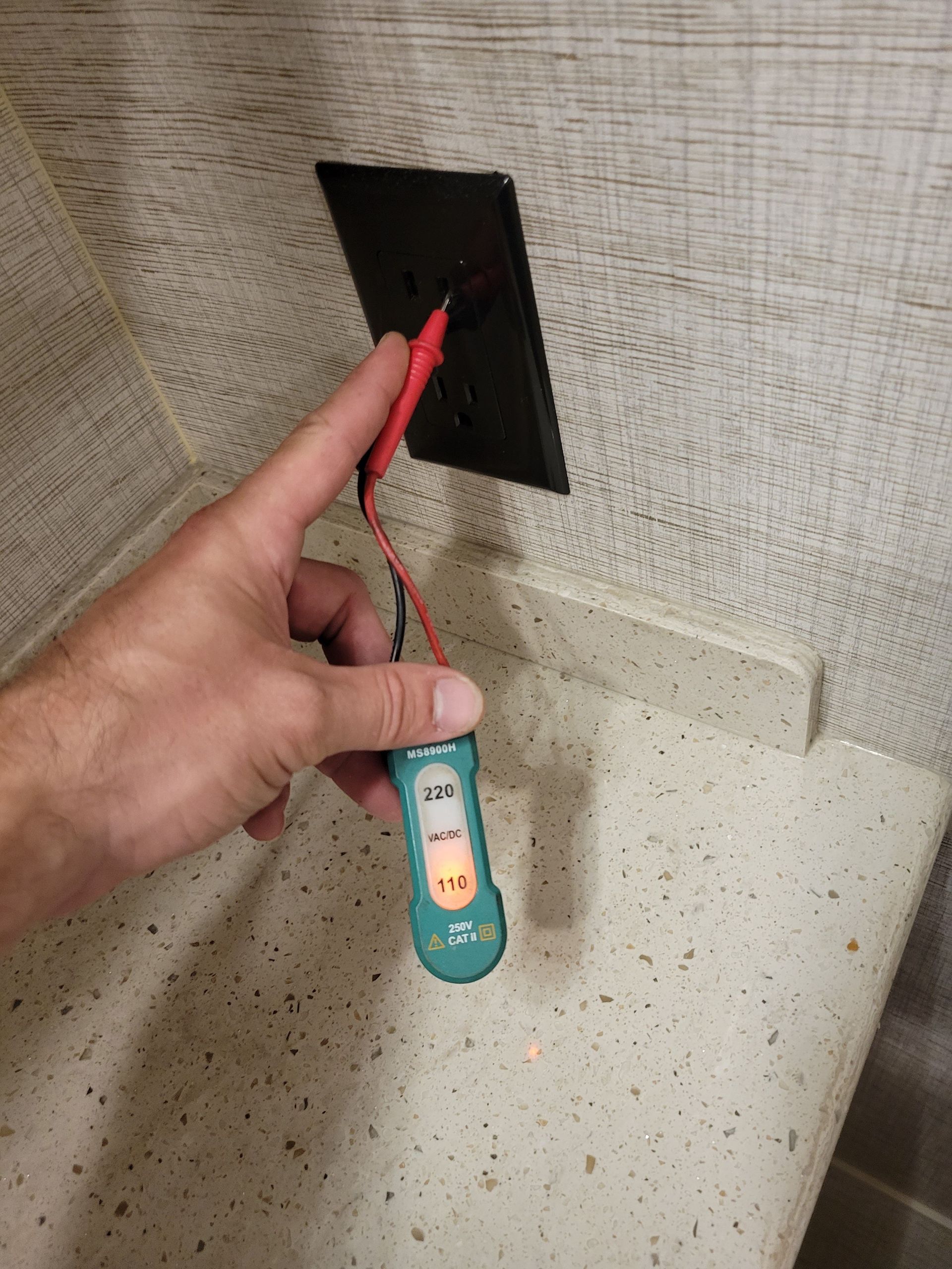 Hand holding a red wire to a wall outlet above a speckled countertop, with a small tester tool attached.