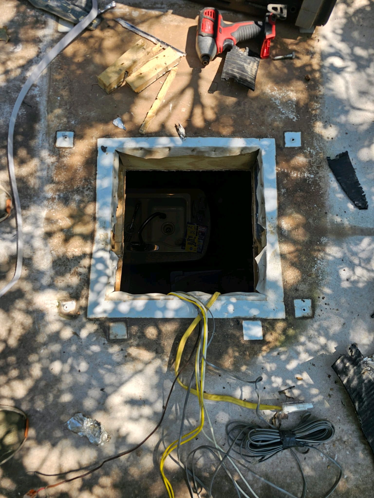 Open square utility hole in pavement with tools, cords, and dirt around the opening