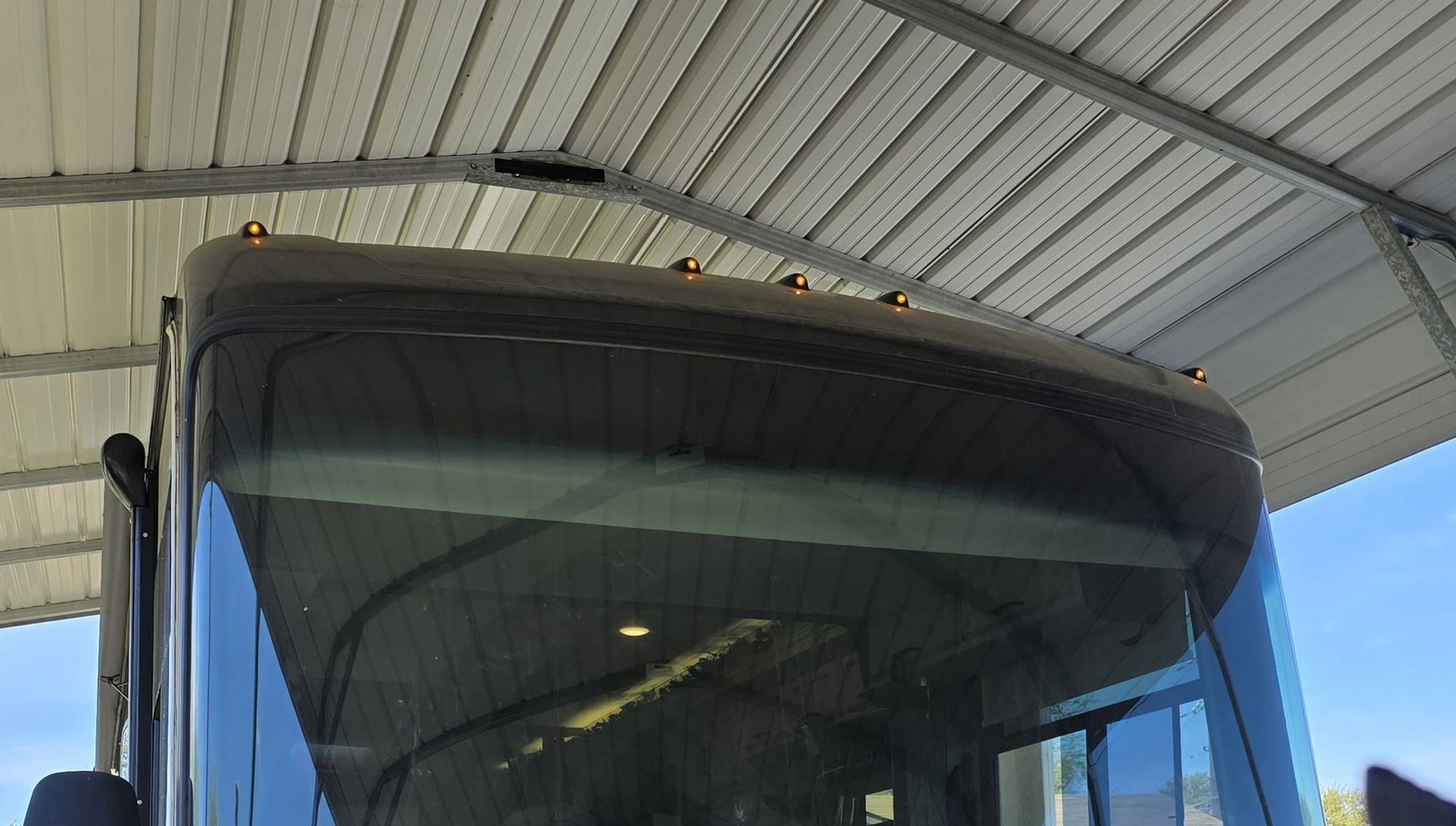 Front of a blue bus under a metal roof, showing the windshield and roof lights.