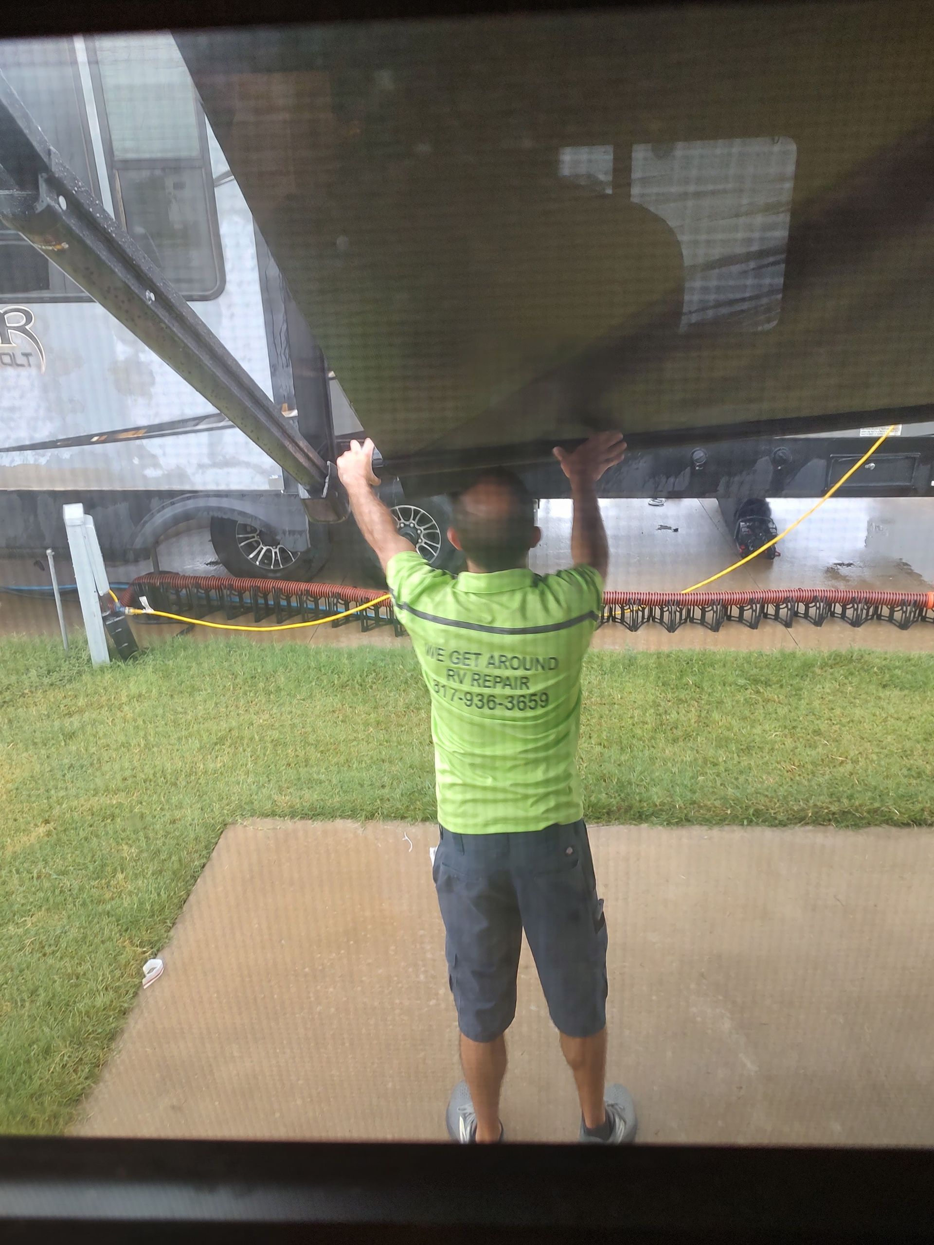 Person in a green shirt and shorts holding up a tarp or canopy in a grassy outdoor area.