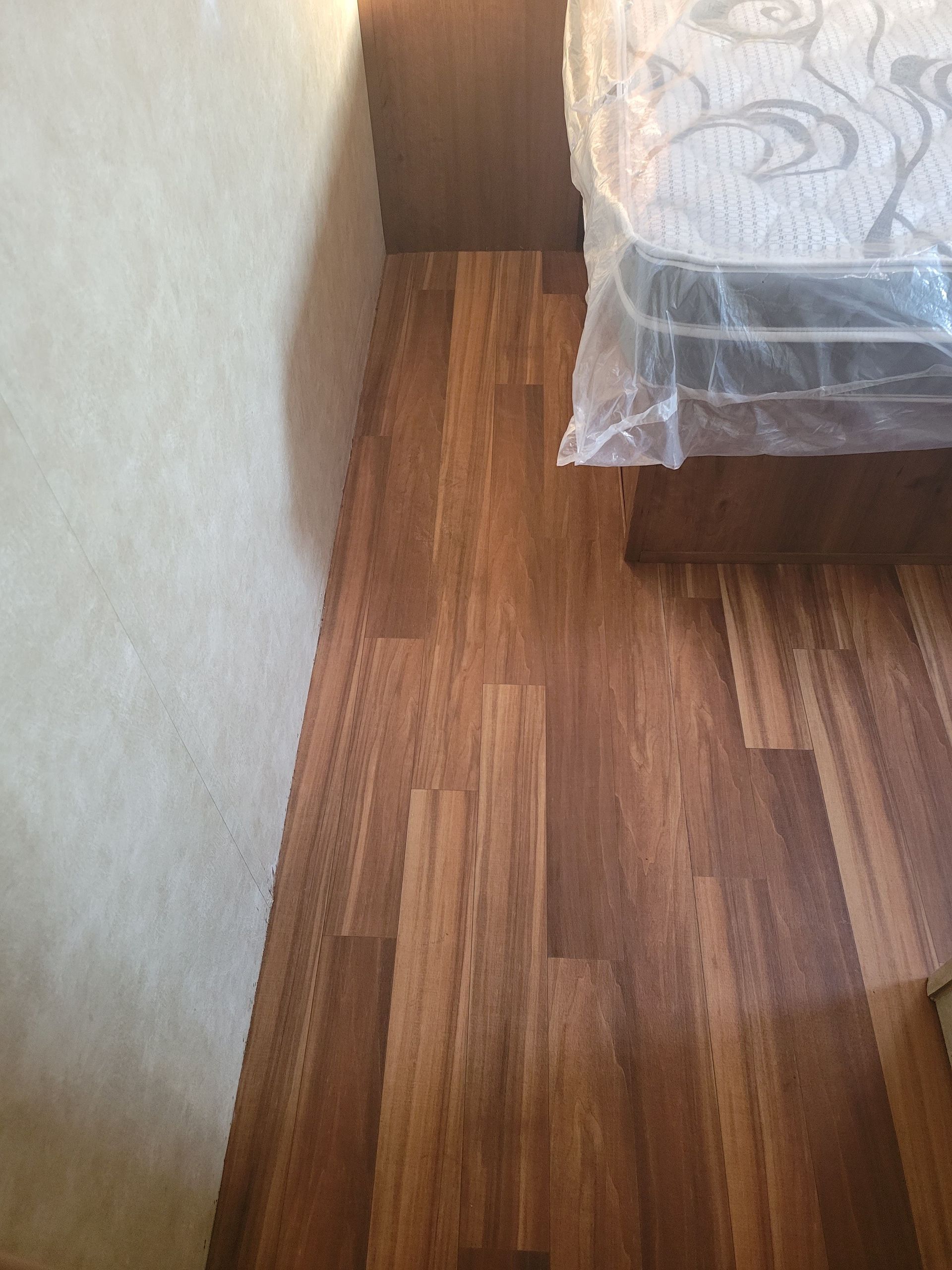 Narrow bedroom gap beside a bed with a plastic-covered mattress and wood flooring.