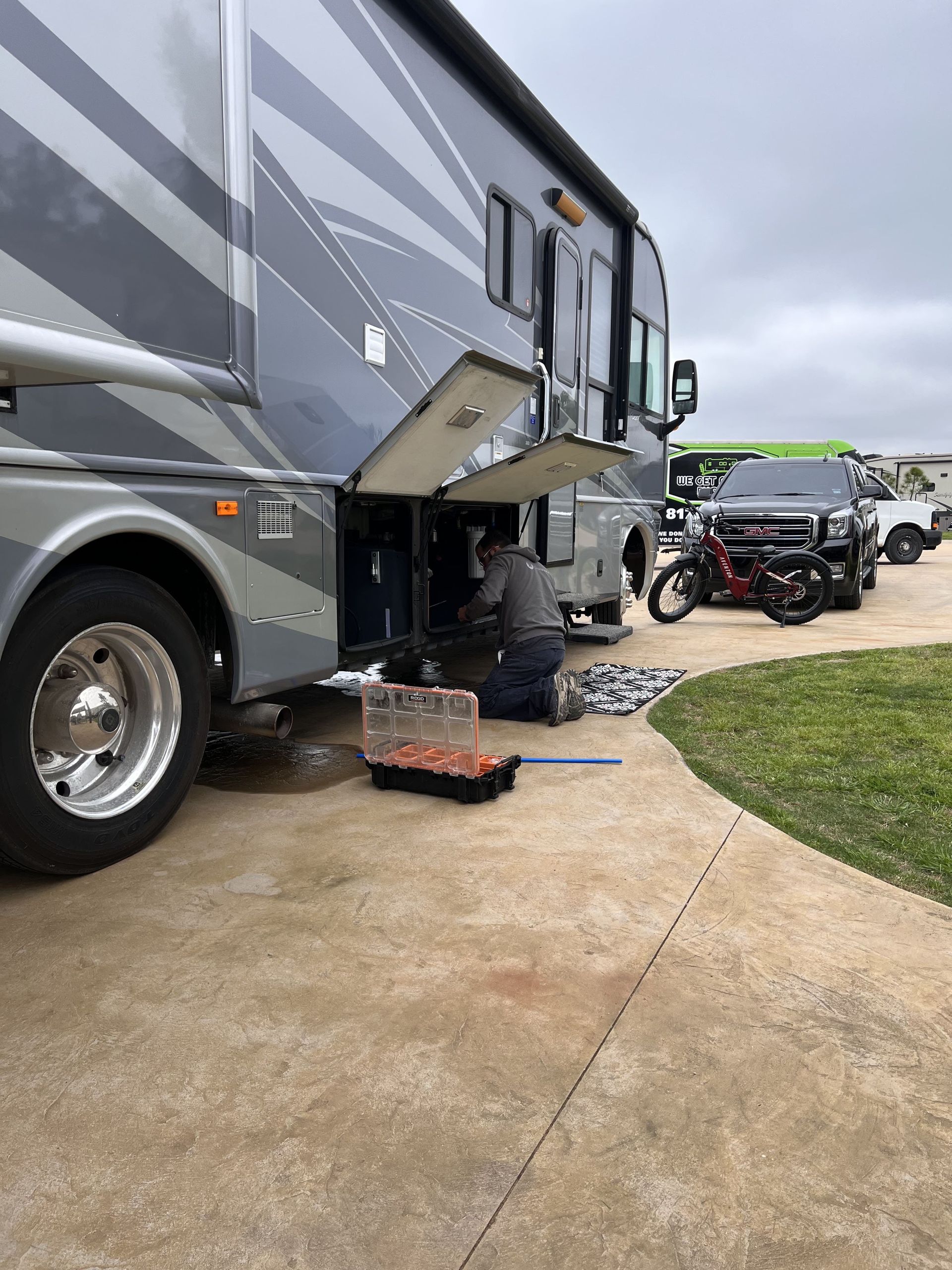 Motorhome parked on a driveway with an open storage compartment and a motorcycle trailer nearby.