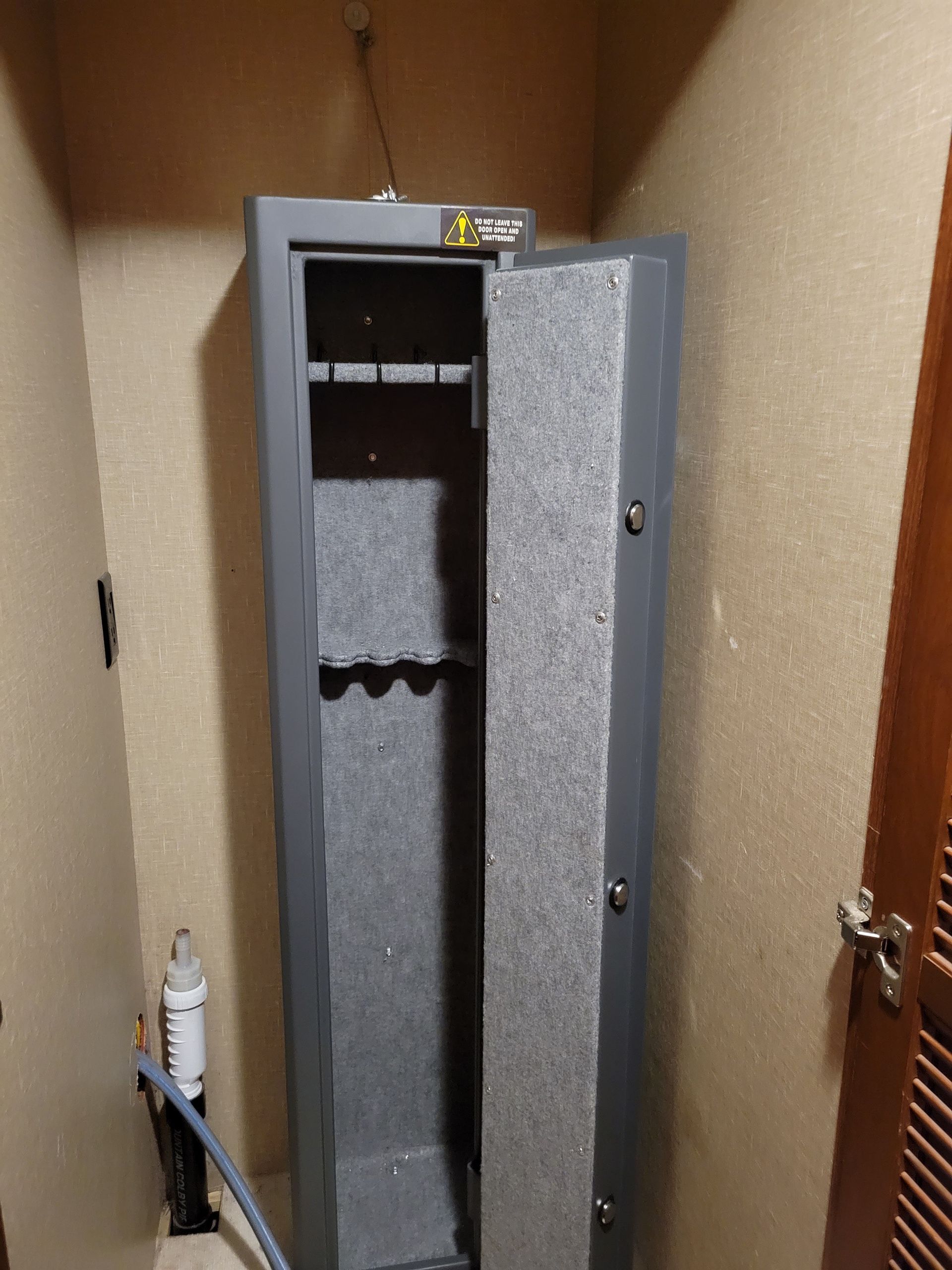 Open gray gun safe in a narrow wooden closet, with shelves and a white pipe beside it