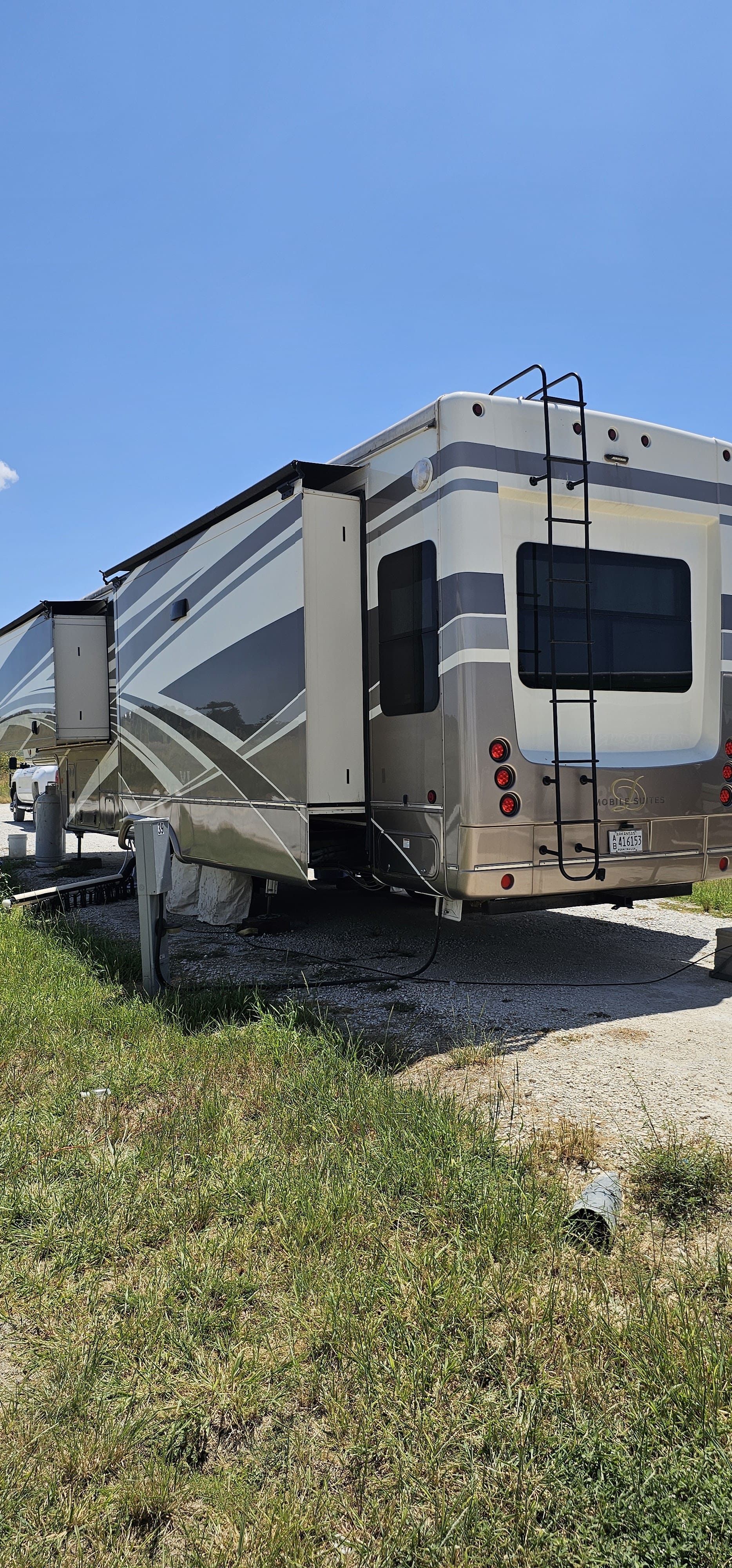 Large white and gray fifth-wheel RV parked on gravel beside grass under a clear blue sky