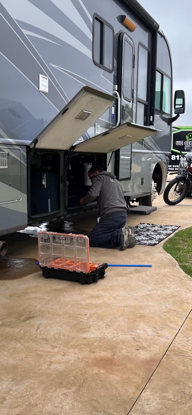 Person crouches under an RV’s open side panel, working on repairs with tools nearby.