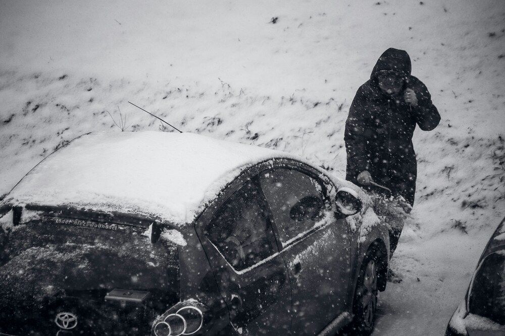 A black-and-white, high-angle shot capturing a person clad in a dark hooded winter jacket clearing snow off a car. The vehicle's roof is buried under a thick blanket of snow, and a snowy hillside serves as the backdrop as fresh snow falls across the frame.