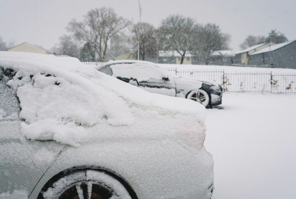 A close-up side perspective of a white car buried under a dense, thick layer of heavy slush and frozen ice. Another packed vehicle sits in the blurry background behind a metal fence, capturing the aftermath of a major freezing weather event.