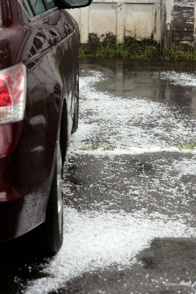 A close-up view of a maroon car parked next to a wet driveway covered in scattered patches of white hail.