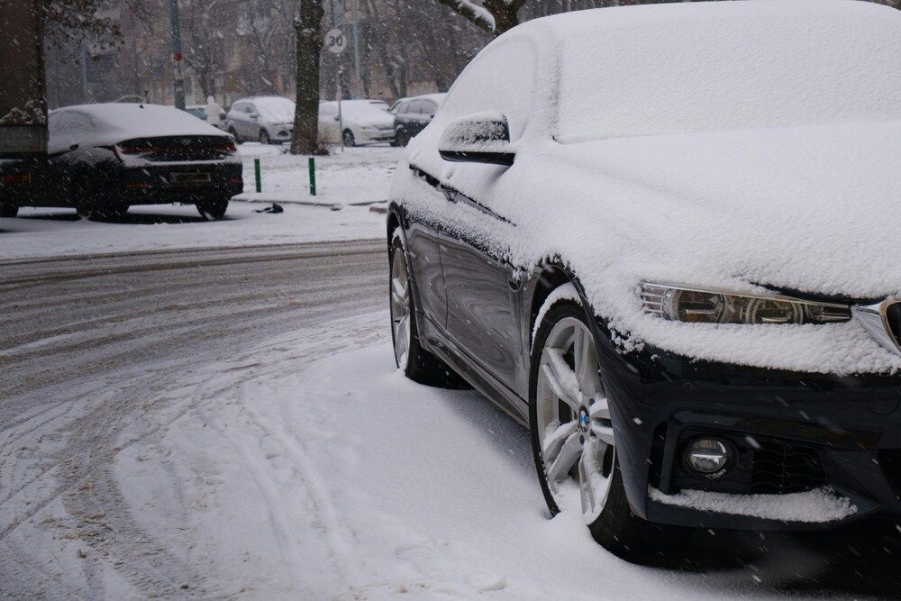 A close-up, angled view of a dark car parked on a snowy city street, with a thick layer of fresh snow covering its hood, windshield, and roof.
