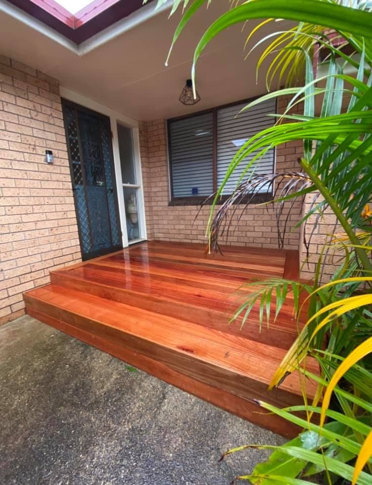 A wooden porch with stairs leading up to a brick house surrounded by plants.
