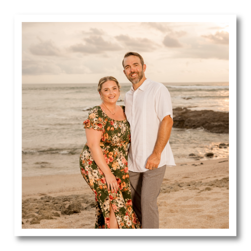 Couple standing on a beach at sunset, smiling and dressed in a floral dress and white shirt.