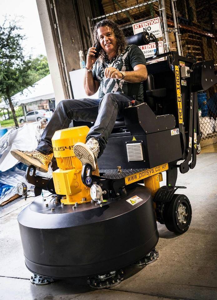 Person riding a yellow floor scrubber in a warehouse, talking on a phone.