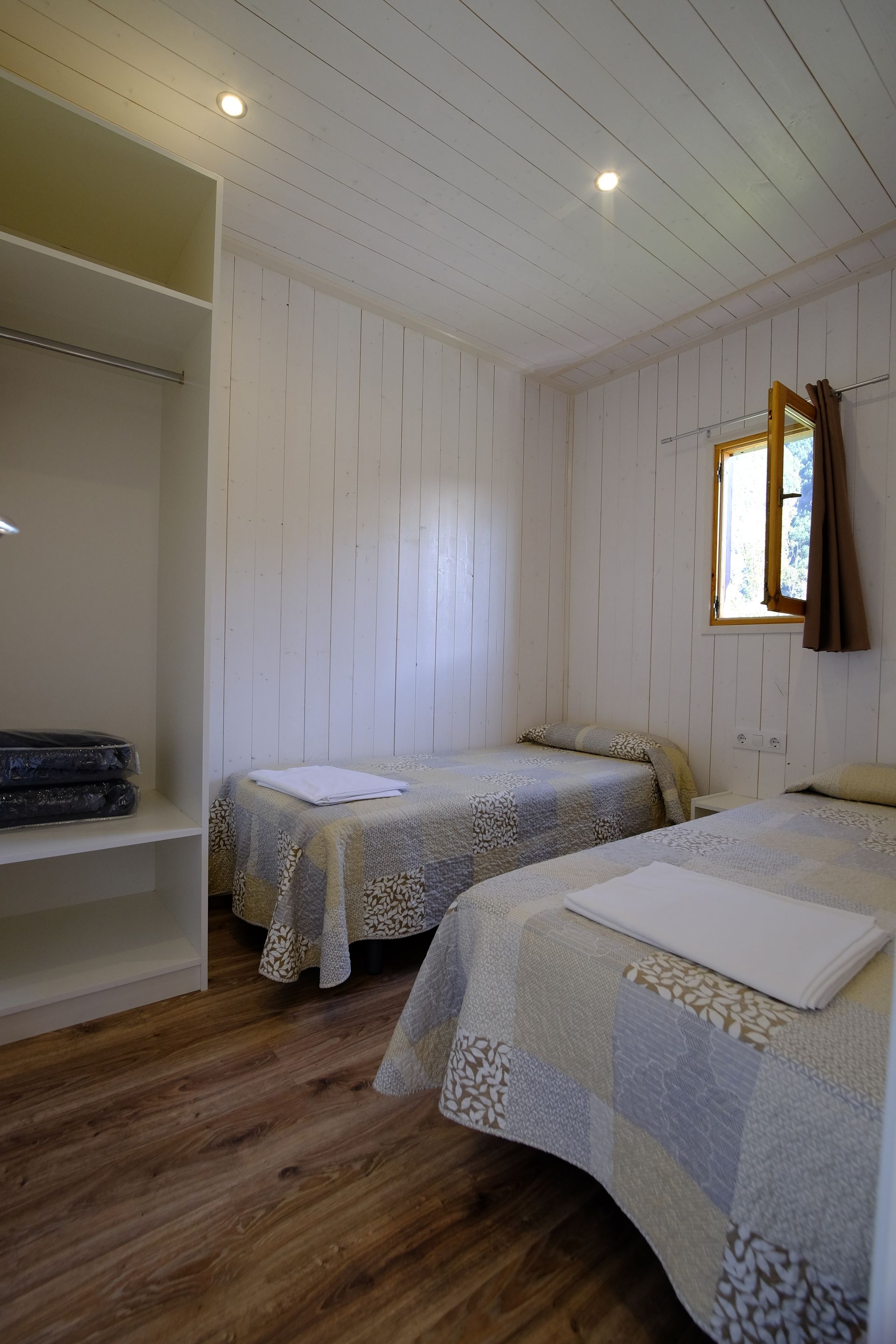 Dormitorio con dos camas individuales, ventana pequeña, armario abierto y suelo de imitación madera.