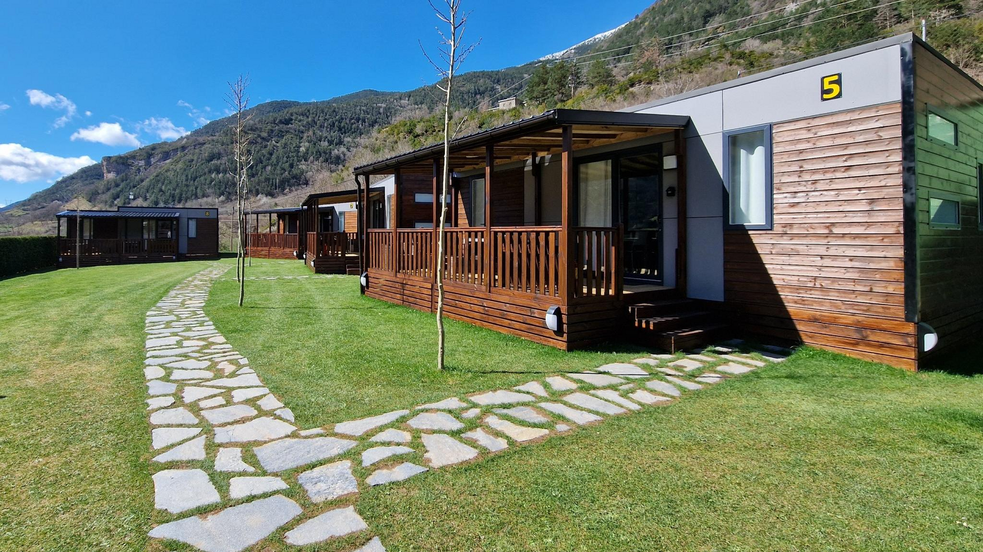 Cabañas de madera con terrazas y sendero de piedra sobre césped verde con vistas a la montaña.