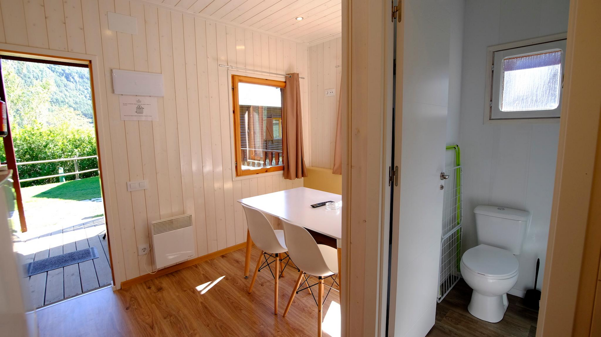 Interior de una cabaña con mesa de comedor, sillas y baño. La puerta abierta ofrece vistas al exterior.