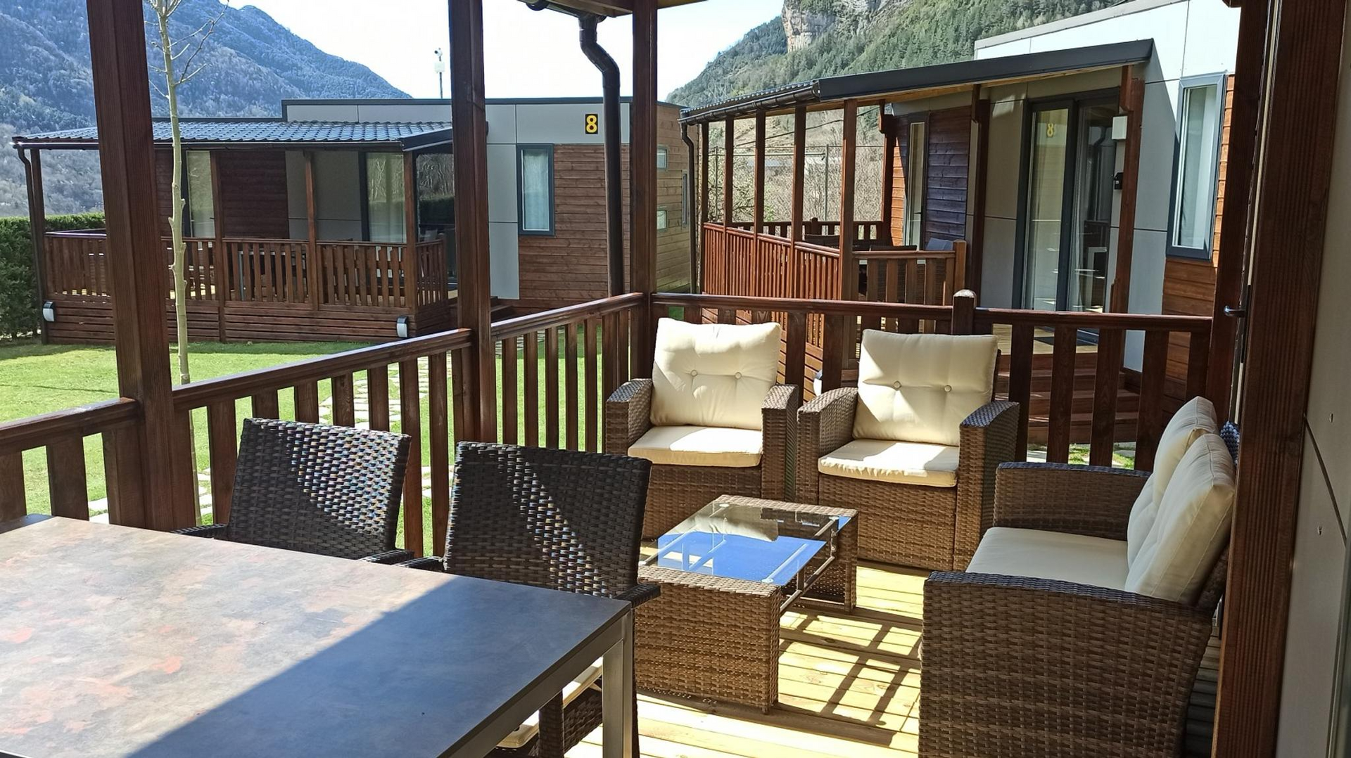 Zona de estar al aire libre con muebles de mimbre y terraza de madera frente a una cabaña, con las montañas de fondo.