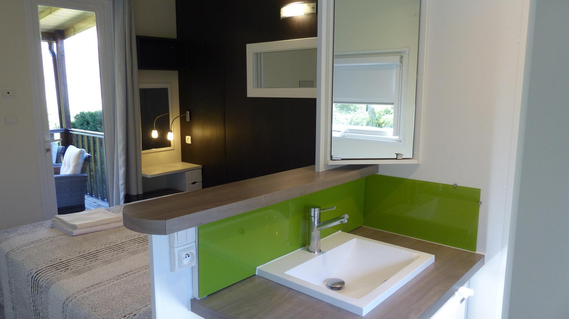 Zona de baño con lavabo y espejo, junto a un dormitorio con cama y balcón.