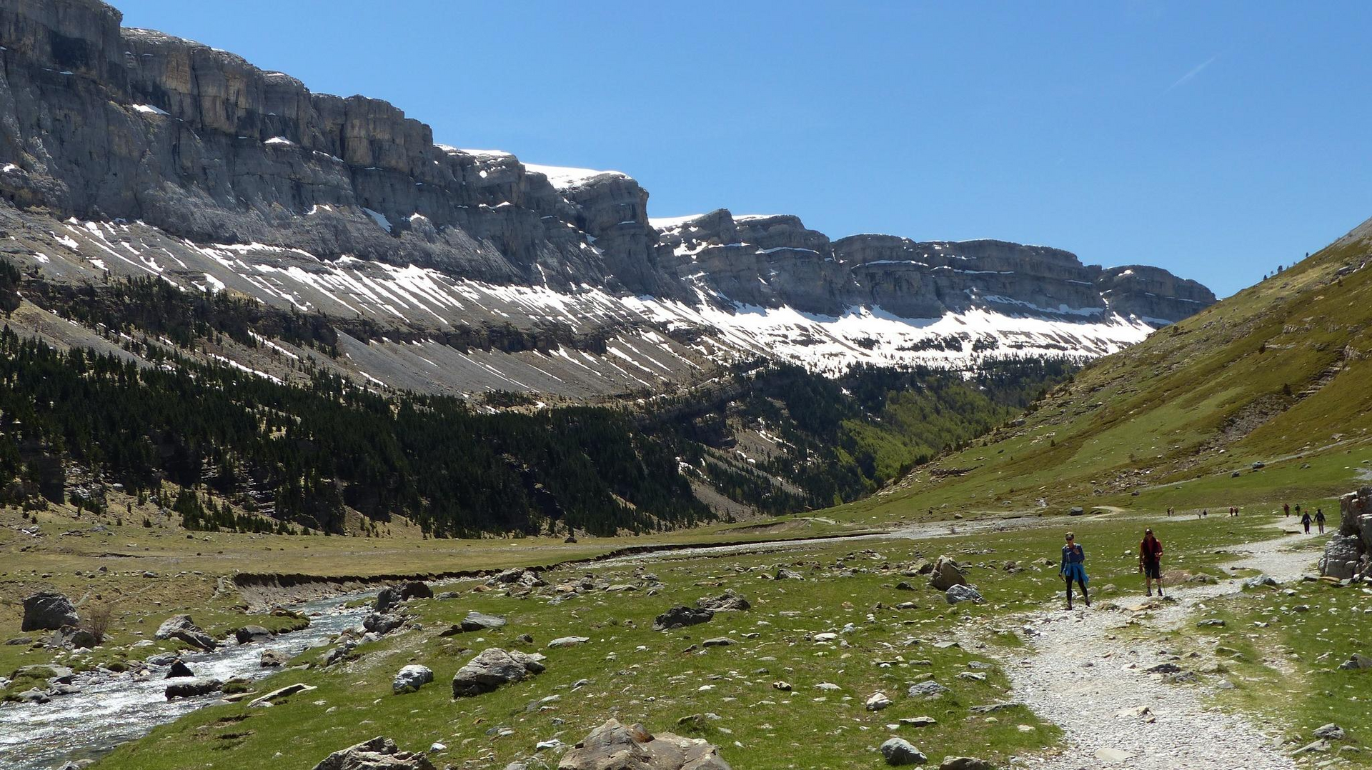 Escena del valle: excursionistas en un sendero, arroyo, hierba verde, acantilados rocosos con manchas de nieve bajo cielo