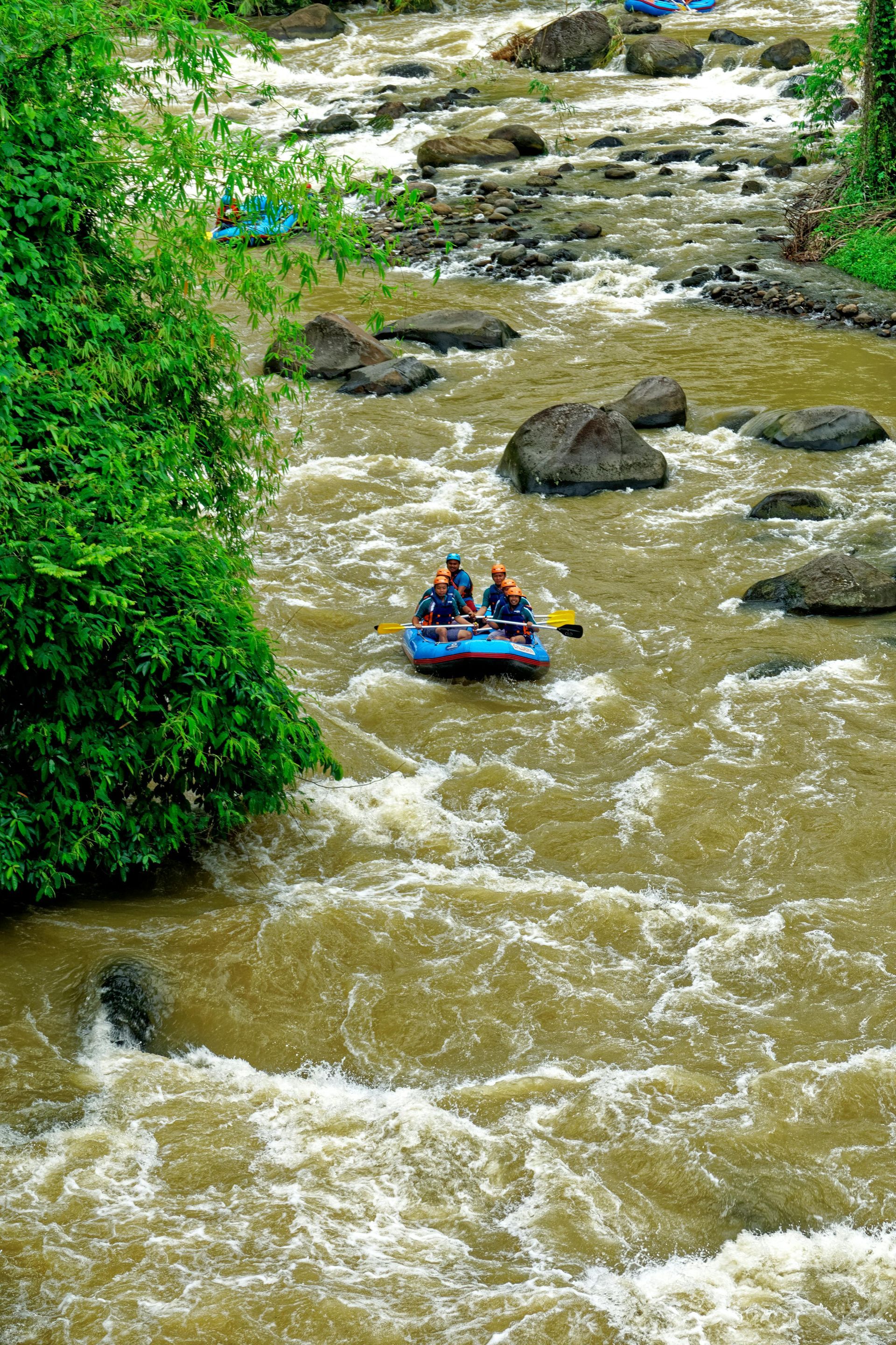Descenso en balsa por un río marrón y caudaloso; dos personas en la balsa, rodeadas de rocas y vegetación verde.
