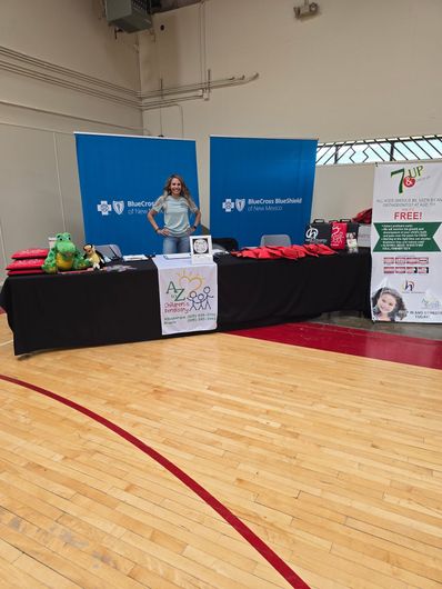 A woman stands behind a table in a gymnasium. The table is covered with promotional materials. A woman stands behind a table in a gymnasium. The table is covered with promotional materials.