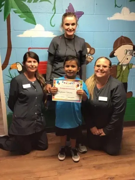A boy holds a certificate with three dental assistants in a colorful clinic; all smile.