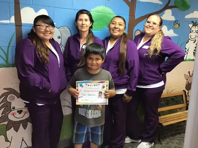 Boy holding certificate with four women in purple uniforms, by a painted wall.