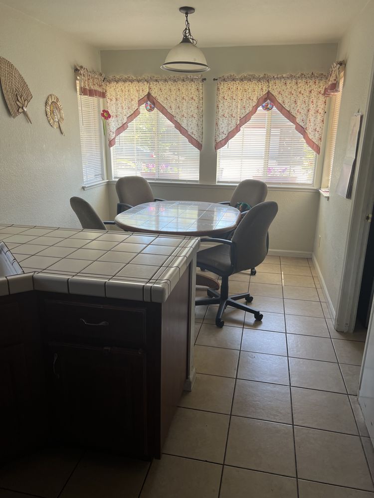 Small dining area with round glass table, four chairs, tiled floor, and sunlit windows with curtains