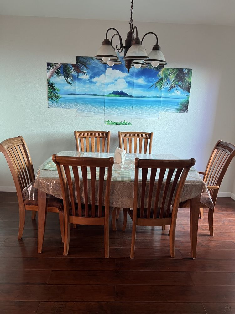 Wooden dining table with six chairs beneath tropical beach wall art and a chandelier.