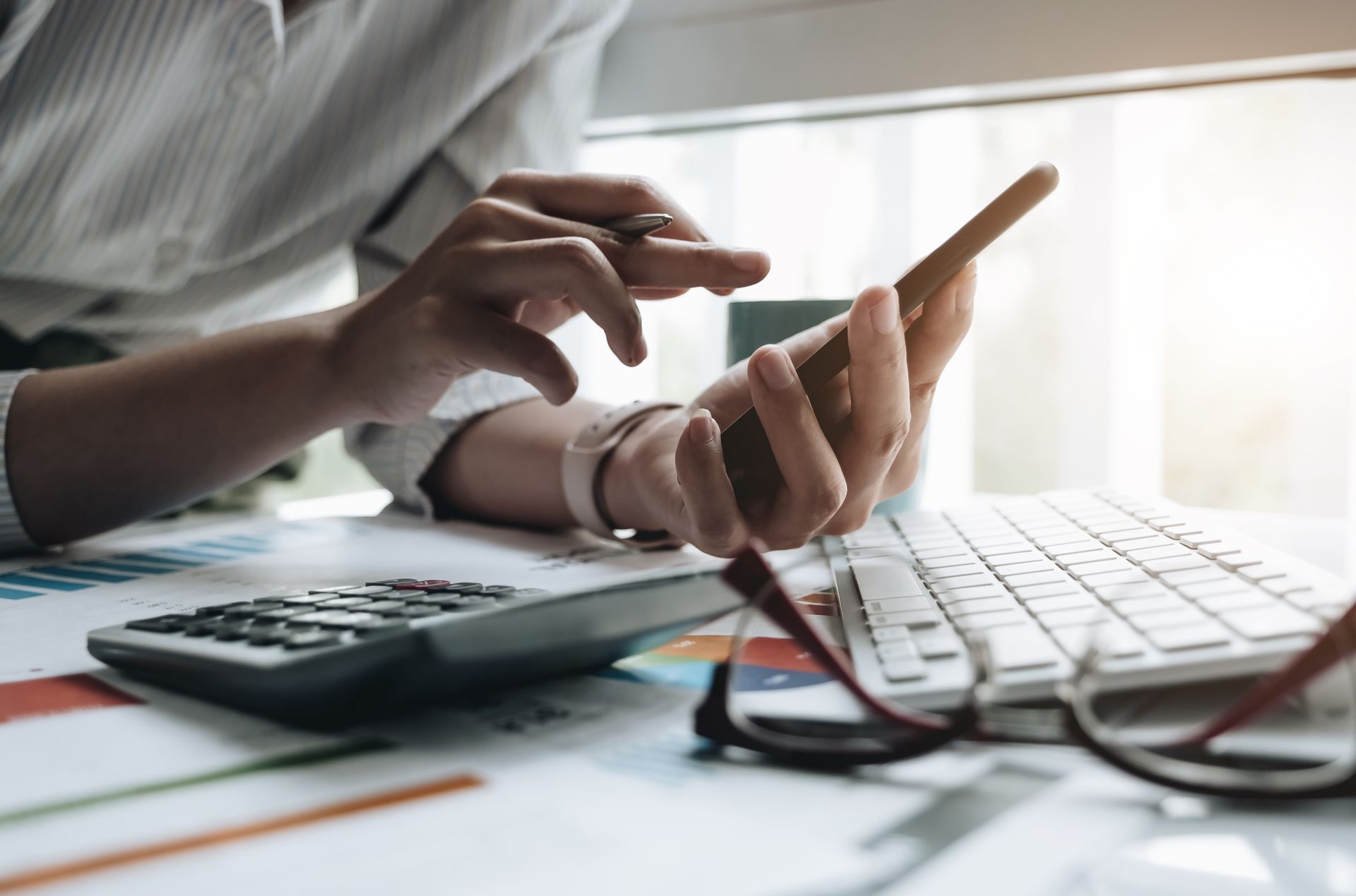 Person using a smartphone at a desk with a calculator, keyboard, and glasses.
