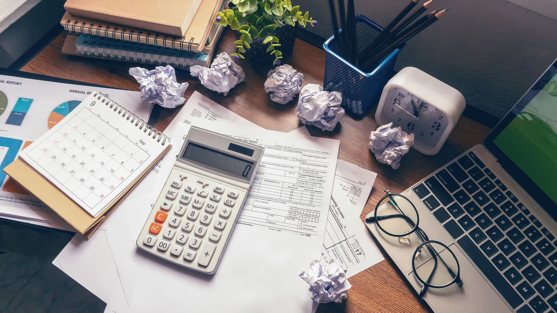 Desk with calculator, laptop, papers, and crumpled balls, suggesting work stress.