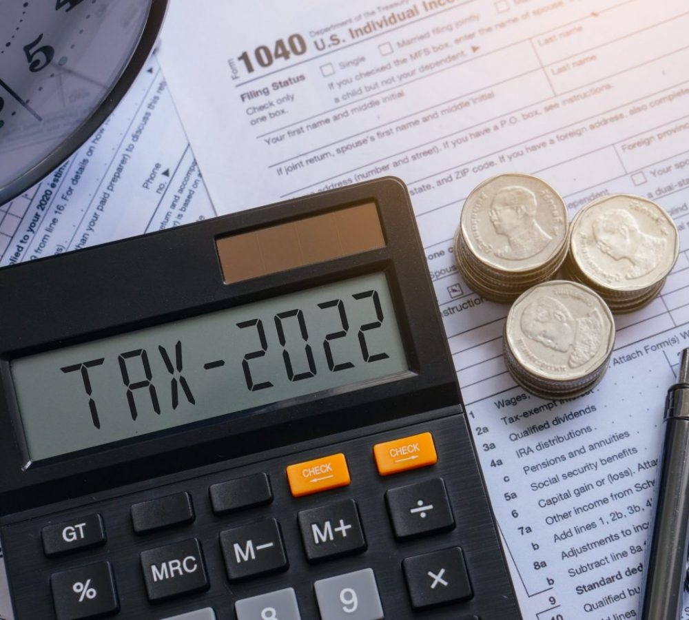Person using a calculator at a desk with a laptop, notebook, phone, and plant. Natural light and green plants visible.