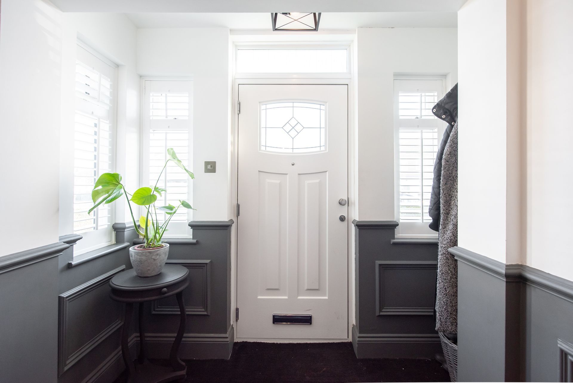 A bright entryway with white walls, a dark gray wainscoting, a closed white door, and a potted plant.