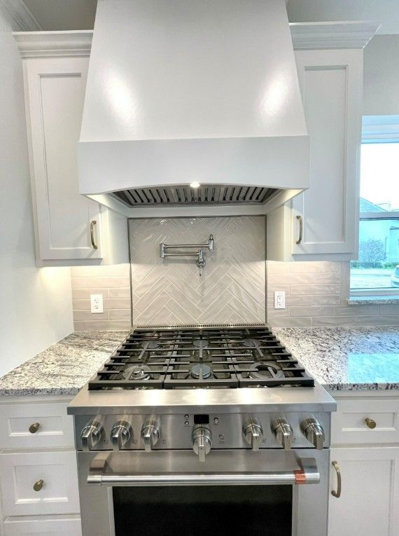 Stainless steel gas range with a white hood and cabinets; backsplash is neutral tile.