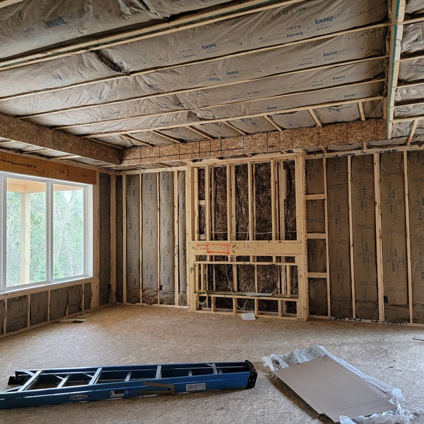 Unfinished room with exposed studs, insulation, and a window; ladder and drywall panel on the floor.