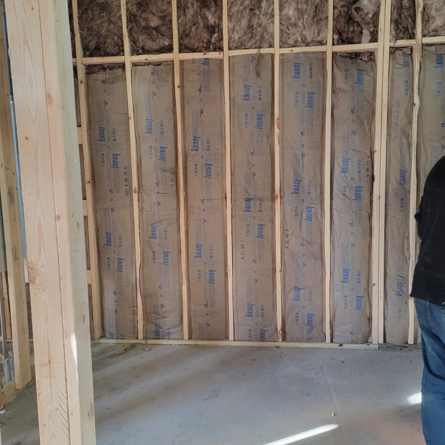 Unfinished interior room with exposed wooden framing, insulation, and a concrete floor; part of a person at right.