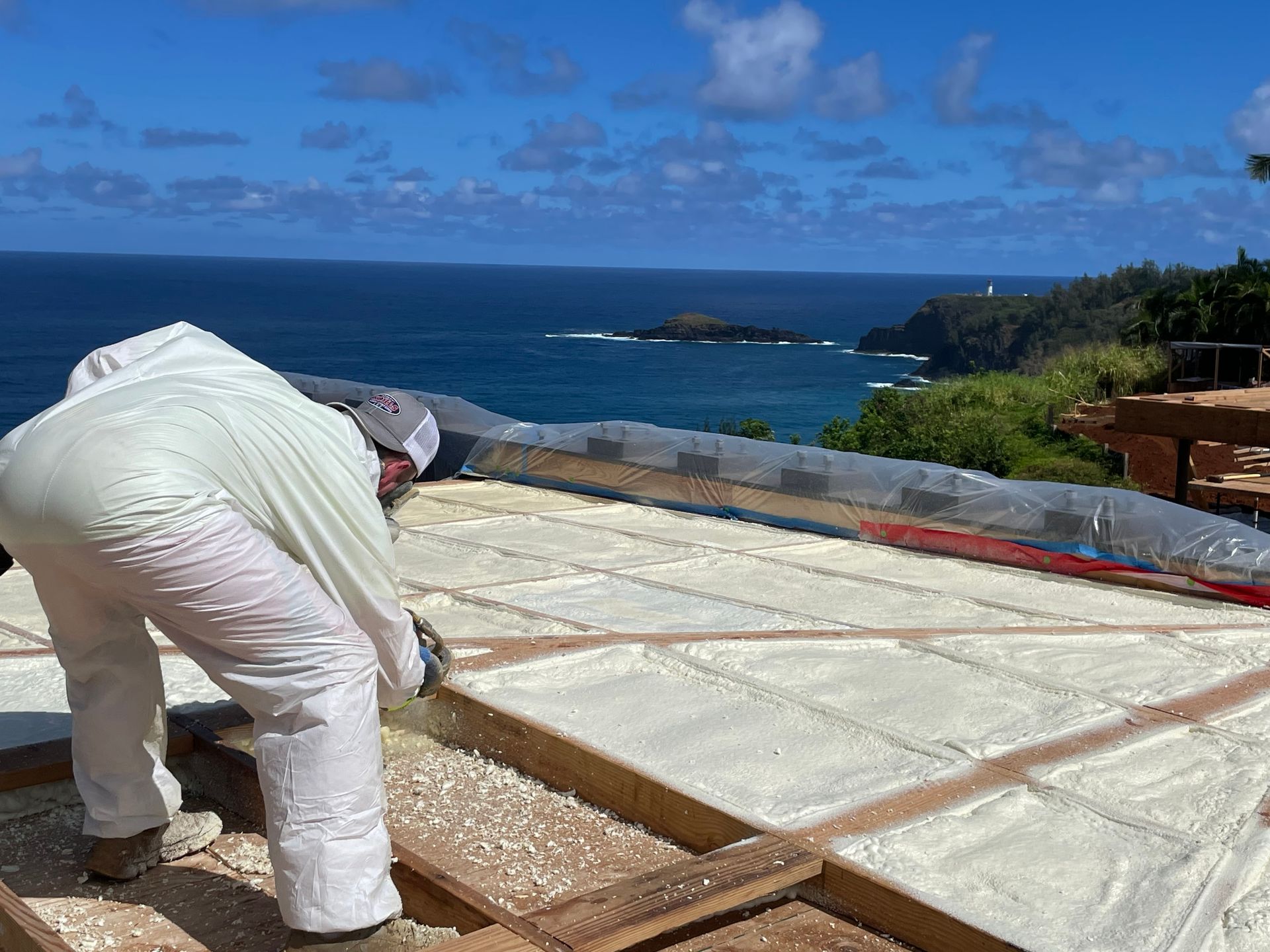 Worker in white suit applying coating on a rooftop with ocean view under a blue sky