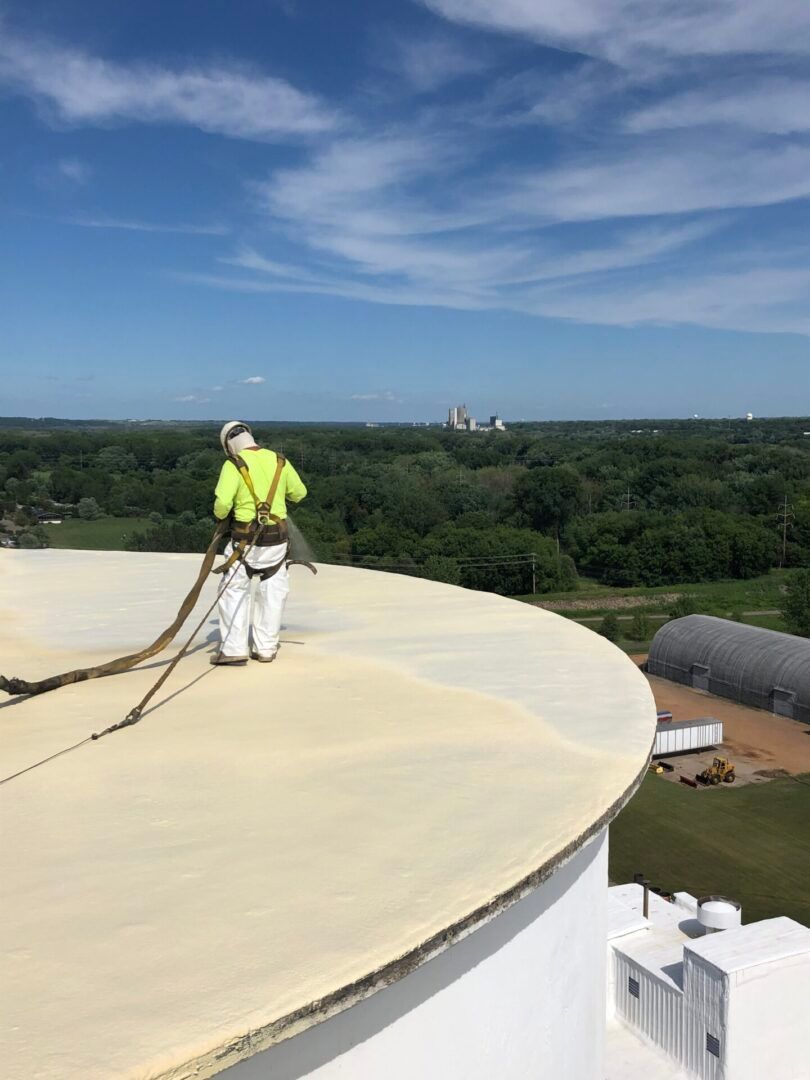 Worker in neon safety gear on a rooftop, applying coating to a large white dome under a blue sky.